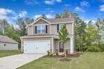 Located at 106 Flue Court in Oxford, this two-story beige house with white trim features a double garage, a concrete driveway, and a lush lawn with young trees and shrubs set against tall trees under a clear blue sky.