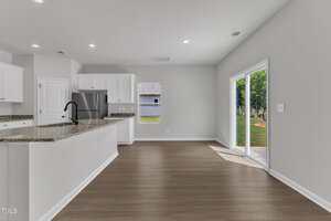 A modern, empty kitchen and dining area with white cabinets, stainless steel appliances, granite countertops, light gray walls, wood-look flooring, a central island, and sliding glass doors leading to a backyard.
