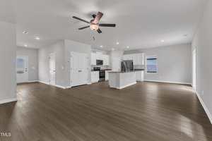 Spacious open-plan kitchen and living area with light gray walls, wood-look flooring, white cabinetry, stainless steel appliances, a ceiling fan, and multiple windows and doors providing natural light.