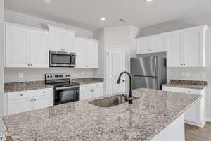 A modern kitchen with white cabinets, stainless steel appliances, granite countertops, an island with a sink, and light gray walls. The space is well-lit with recessed ceiling lights.