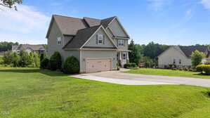 Two-story suburban house with gray siding and an attached two-car garage. The home is set on a large, well-maintained lawn with a concrete driveway, surrounded by other houses and greenery under a blue sky.