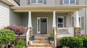 A front porch of a house with stone steps, white pillars, and a gray front door. Potted plants and shrubs line the steps, and the house exterior features gray siding and double windows.
