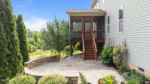 A stone patio with surrounding landscaping is shown next to a house with beige siding. Wooden stairs lead up to a screened porch. Tall shrubs and trees are visible in the background.
