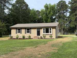 A single-story beige brick house at 2253 Nc Highway 96, Franklinton, with a gable roof, black front door, and three windows. The grassy yard features small shrubs and trees in the background beneath a partly cloudy sky.