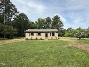 A single-story, tan brick house with a dark roof sits on a large, well-maintained lawn at 2253 NC Highway 96 in Franklinton. There are a few small shrubs in front of the house, and trees border the property in the background.