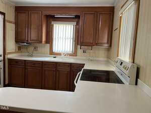 A kitchen with wooden cabinets, white countertops, an electric stove, a double sink under a window with vertical blinds, and neutral-colored walls. Natural light enters through the window above the sink.