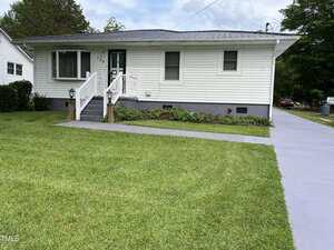 Charming single-story white house at 129 S Bullock Street, Henderson features a metal roof, front porch with steps and handrails, gray concrete driveway and walkway, green lawn, foundation bushes, and trees in the background.