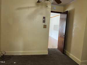 A small room with beige walls and carpet, an open wooden door, a ceiling fan above, and some visible wall switches and outlets. Cables are on the floor in the corner, and daylight enters from the adjacent room.