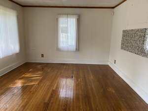An empty room with hardwood floors, two windows with vertical blinds, beige walls, a rectangular vent on the floor, and a section of wall with patterned wallpaper. Sunlight streams through the blinds onto the floor.