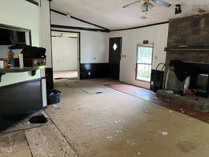 A living room with partially removed flooring, visible subfloor, and scattered debris. There is a stone fireplace, a ceiling fan, and an open doorway to another room. A black door and window let in natural light.