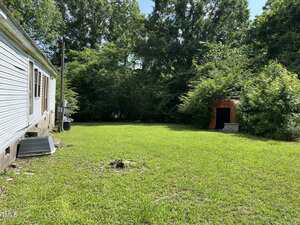 A grassy backyard with a white-sided house on the left, an air conditioning unit, and a small, rusted metal shed surrounded by dense trees and bushes under a clear blue sky.