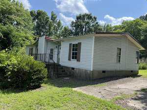 A single-story white house with brown shutters, a metal roof, and a small set of front steps at 2511 Brogden Road in Creedmoor, surrounded by grass, trees, and bushes under a partly cloudy sky.