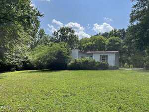 A small white mobile home is surrounded by green bushes and trees, sitting on a large grassy yard under a partly cloudy blue sky.