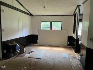 A mostly empty room with white and black walls, a large window at the far end, and a sheet of white material leaning against the window. Boxes and miscellaneous items are in one corner, and the floor is partially unfinished.