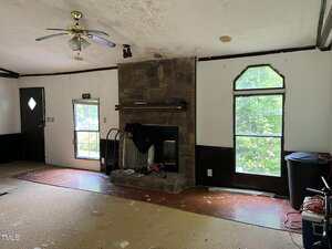 A living room with an unfinished floor, a stone fireplace in the center, a ceiling fan, and two tall windows. There are various items and some debris scattered around the room.