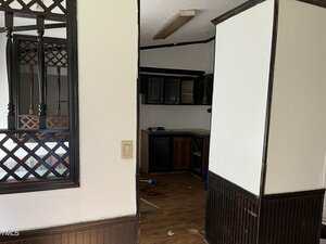 A view into a small, dimly lit kitchen with dark cabinets and wood flooring. The kitchen is partially visible through an open wall with wood trim and decorative lattice on the left side.