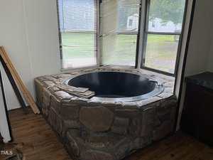 A round black bathtub set in a stone surround is positioned in a corner next to large windows with partially open blinds. Wood flooring surrounds the tub, and a few wooden boards rest nearby.
