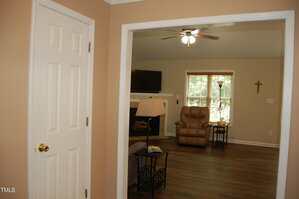 View into a living room with beige walls, a ceiling fan, a brown recliner, a floor lamp, a wall-mounted TV above a fireplace, a window with blinds, and a wooden cross on the wall.