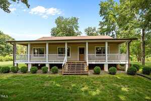 A charming one-story house at 3044 Rock Springs Church Road in Creedmoor features a large covered front porch with white railings, beige siding, a brown roof, and wooden steps. Lush green lawn, shrubs, and trees complete the inviting setting.