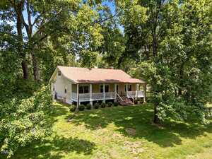 A single-story house with a large covered front porch and stairs, situated on a grassy lawn and surrounded by tall trees under a clear blue sky.