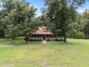 A tan, two-story house with a covered front porch sits behind two large trees in a spacious grassy yard surrounded by woods. A walkway leads from the porch to the foreground.