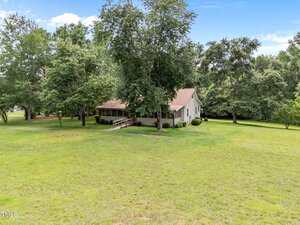 A single-story house with a covered front porch and ramp sits surrounded by a large grassy yard and mature trees under a blue sky with scattered clouds.