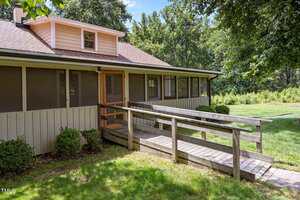 A single-story house with a ramp leading to a screened front porch, surrounded by grass, bushes, and trees under a sunny sky.