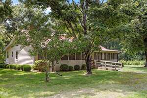A single-story house with beige siding sits among tall trees. The house has a large screened-in porch, a wooden ramp leading to the entrance, and well-maintained bushes along the side. Grass covers the yard.