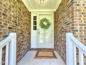 Brick front porch with a white railing, a tan doormat, and a glass front door featuring a green wreath. A light fixture hangs from the white ceiling above the entryway.