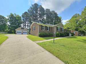 Single-story brick house with white trim, a well-kept lawn, and a wide driveway leading to a detached two-car garage, surrounded by green trees under a partly cloudy sky.