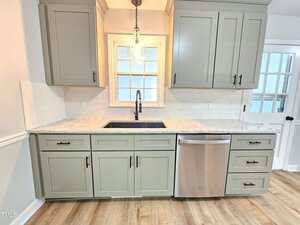 A modern kitchen features light gray cabinets, a granite countertop, a stainless steel dishwasher, a farmhouse-style sink with a gooseneck faucet, a window with divided panes, and wood-look flooring.