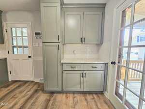 A kitchen corner with light green cabinets, white subway tile backsplash, a marble countertop, and wood flooring. A glass door leads outside, and a white door is visible on the left wall.