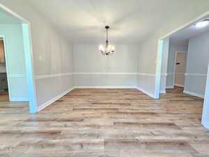 A bright, empty dining room with light-colored walls, wood flooring, white trim, and a black chandelier. Open doorways lead to other rooms, one with visible kitchen cabinets and another with a hallway light.
