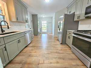 A modern kitchen with green cabinets, stainless steel appliances, light countertops, wood-style flooring, and a view of double doors leading to a bright, sunlit room at the far end.