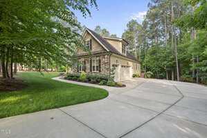 A two-story house with stone and siding exterior, white garage doors, and landscaped front yard, surrounded by tall trees and a wide concrete driveway curving to the entrance.