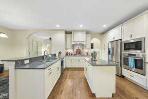 Spacious kitchen with white cabinets, stainless steel appliances, granite countertops, a center island with a vase of flowers, wood flooring, and recessed lighting. An archway leads to another room.