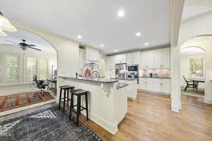 A spacious kitchen with white cabinets, stainless steel appliances, granite countertops, and a breakfast bar with two black stools. Hardwood floors and multiple archways open to adjacent rooms with natural light.