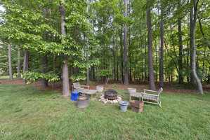 A wooded backyard area with tall trees, a fire pit surrounded by benches and chairs, and various containers and a cardboard box on the grass. The setting is shaded and the ground is covered with green grass.
