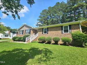 Single-story brick house with white trim, black shutters, and a front porch with white railings. The front yard has neatly trimmed bushes and green grass. Tall trees are visible behind the house under a blue sky.