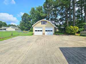 A two-car garage with a second-story window sits at the end of a wide concrete driveway, surrounded by green grass, trees, and a chain-link fence on the left side.