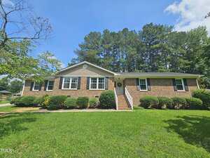 A charming one-story brick house on 112 Saddletree Road in Oxford, with dark shutters, white trim, and a small set of front steps. The grassy yard features trimmed bushes and trees, while tall pines stand beneath a clear blue sky.