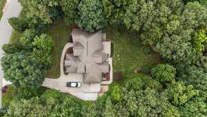 Aerial view of a two-story house with a gray roof, surrounded by dense green trees and a well-maintained lawn; a black and white vehicle is parked in the driveway next to the house.