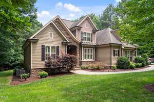 A two-story suburban house with beige siding, dark shutters, stone accents, and a gabled roof sits beside a curved driveway, surrounded by green grass, shrubs, and mature trees on a sunny day.