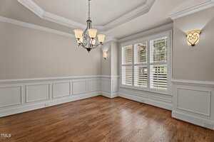 An empty dining room with hardwood floors, beige walls with white wainscoting, a tray ceiling, a chandelier, two wall sconces, and three large windows with white shutters.