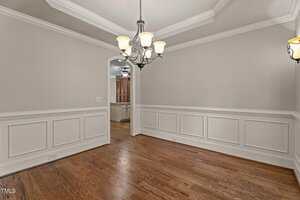 An empty dining room with light beige walls, white wainscoting, hardwood floors, a tray ceiling, and a central chandelier; an open doorway leads to another room with similar decor.