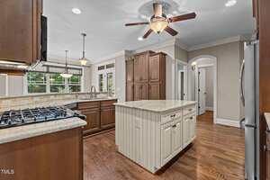 A spacious kitchen with wooden cabinets, a central island with drawers, stainless steel appliances, a ceiling fan with light, and hardwood floors. Large windows provide an outdoor view and natural light.