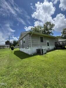 A single-story house with gray siding and white trim sits on a grassy lot. An outdoor air conditioning unit is next to the house, and a wooden deck is visible on the right. The sky above is partly cloudy.