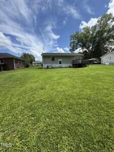 A single-story house with gray siding and a wooden deck in the backyard, situated on a large, well-maintained grassy lawn under a partly cloudy sky. Neighboring houses are visible on either side.