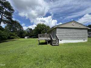 A single-story house with gray siding and a small wooden deck sits on a large grassy lawn. Trees and a small shed are visible in the background under a partly cloudy sky.