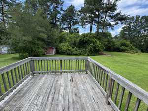 A weathered wooden deck with a railing overlooks a grassy backyard bordered by trees and bushes. A small red shed is visible near the tree line on the left side. The sky is partly cloudy.
